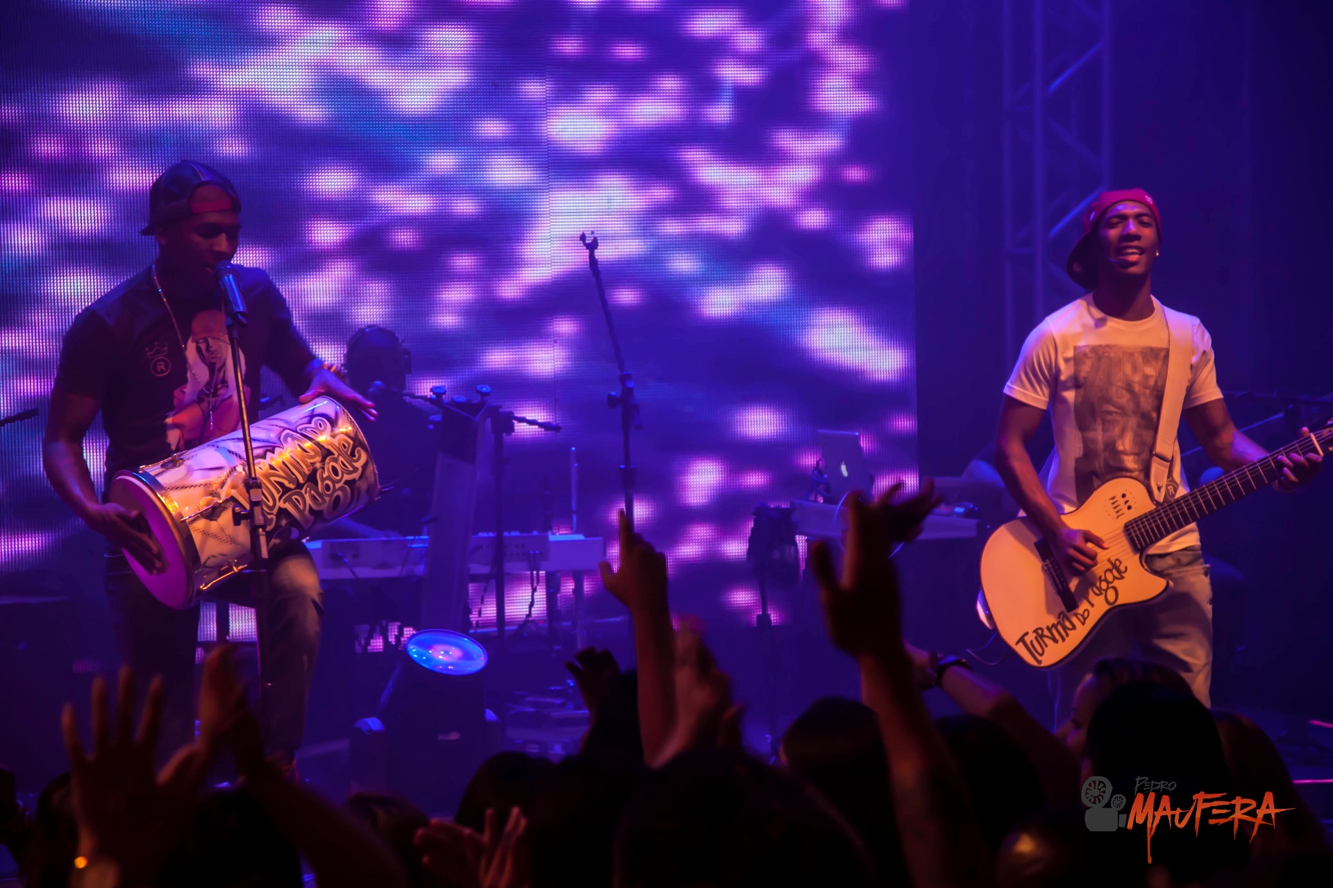 Percussionista e guitarrista sob luzes roxas com plateia